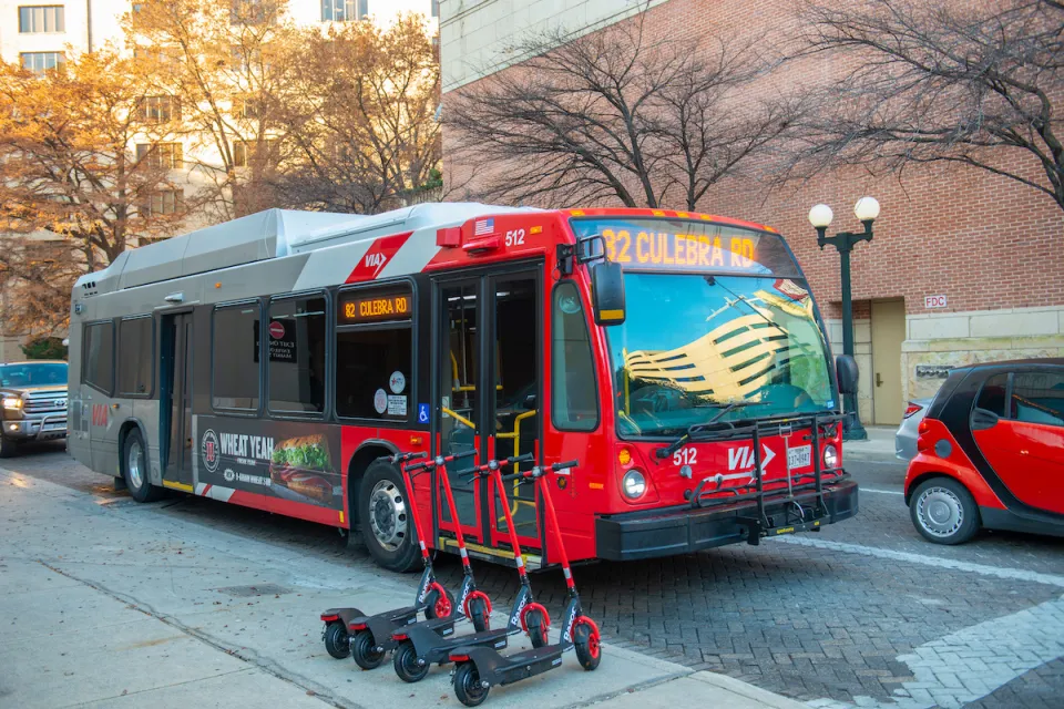 Red public bus on street in San Antonio, Texas with line of four red e-scooters on sidewalk.